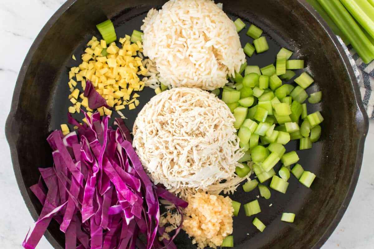 overhead shot of each ingredient seperated into their own sections in a cast iron skillet: white rice, sliced red cabbage, shredded chicken, fresh ginger, celery sticks, minced garlic, soy sauce and sesame oil