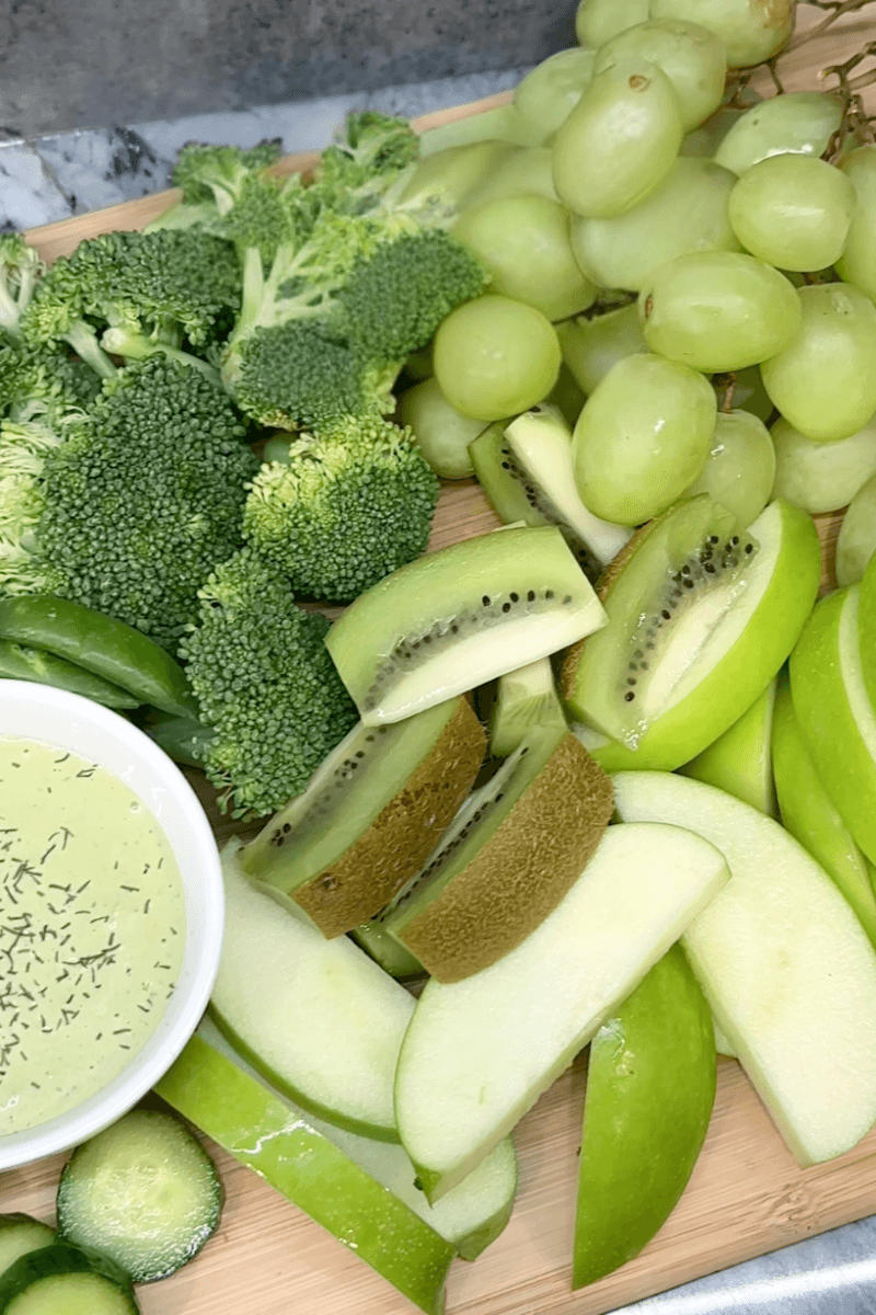 Close up of board on kiwi, apples, grapes, broccoli, cucumbers and dip