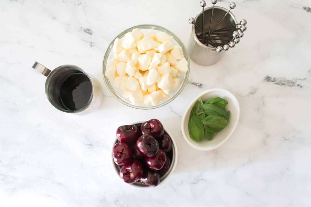 A flat lay overhead shot on a white marble countertop showcasing the recipe ingredients: a bowl of pitted dark sweet cherries, a bowl of fresh mozzarella pearls, a small dish of fresh green basil leaves, a small metal pitcher of balsamic glaze, and a metal cup holding cocktail skewers.