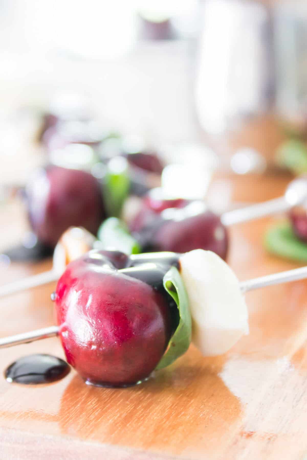 A close-up shot of bite-sized sweet cherry caprese skewers arranged beautifully on a dark slate serving board, showing the layers of dark red pitted cherries, fresh green basil leaves, and white mozzarella pearls drizzled with a rich balsamic glaze.