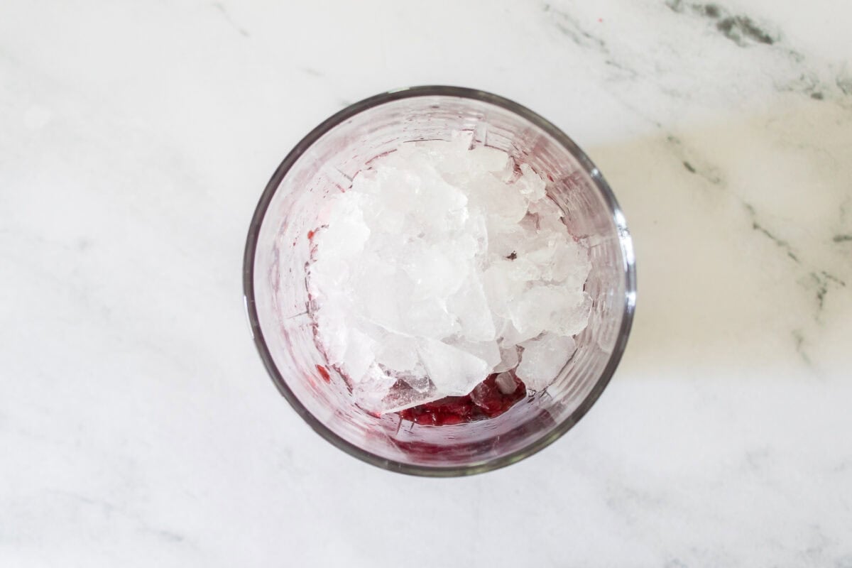 A close-up shot of crushed ice being poured from a small stainless steel pitcher into a lowball glass already filled with muddled dark red cherries, set on a bright marble countertop.