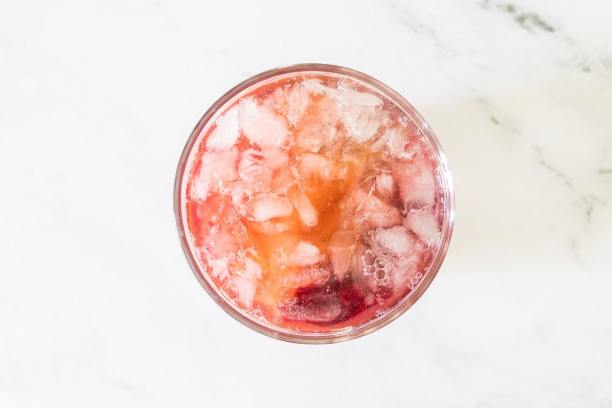 A close-up shot of bright yellow mango nectar being poured from a glass measuring cup into a lowball glass filled with crushed ice and muddled dark cherries, showing the ingredients beginning to mix on a marble countertop.