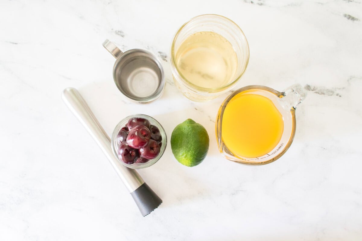 A flat lay ingredient shot on a marble countertop showing a cocktail muddler next to a small glass bowl of fresh cherries, a whole lime, a measuring cup of bright yellow mango nectar, a jar of simple syrup, and a small metal jigger.