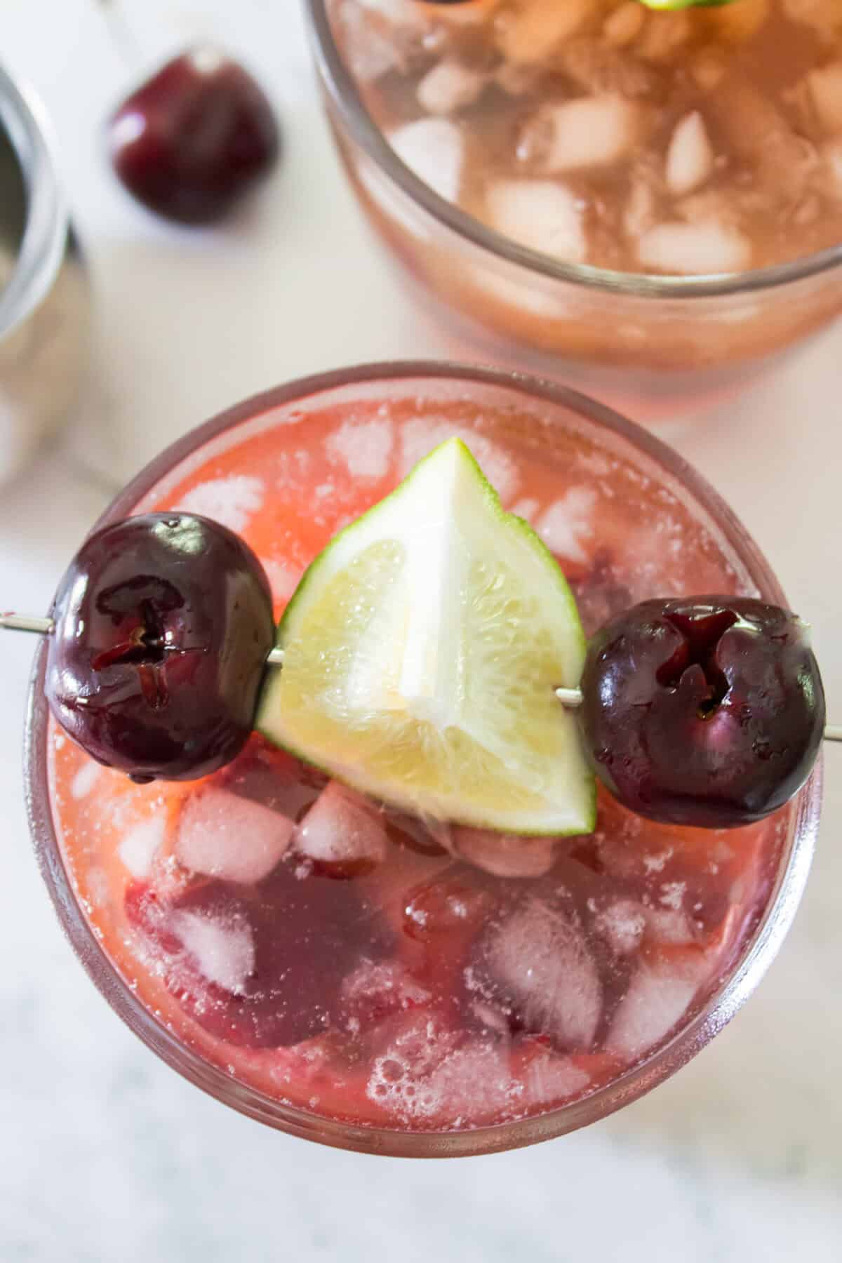 An overhead, close-up shot of a refreshing cherry mango margarita filled with crushed ice, showcasing its vibrant reddish-pink color and garnished with a cocktail skewer featuring two fresh dark sweet cherries and a green lime wedge.