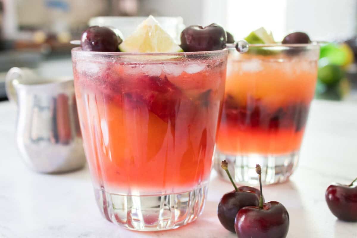 A close-up, side-angle shot of a cherry mango margarita in a lowball glass filled with crushed ice, showcasing a beautiful ombre effect as the dark red muddled cherry juices at the bottom naturally bleed up into the vibrant peach-colored mango nectar layer.