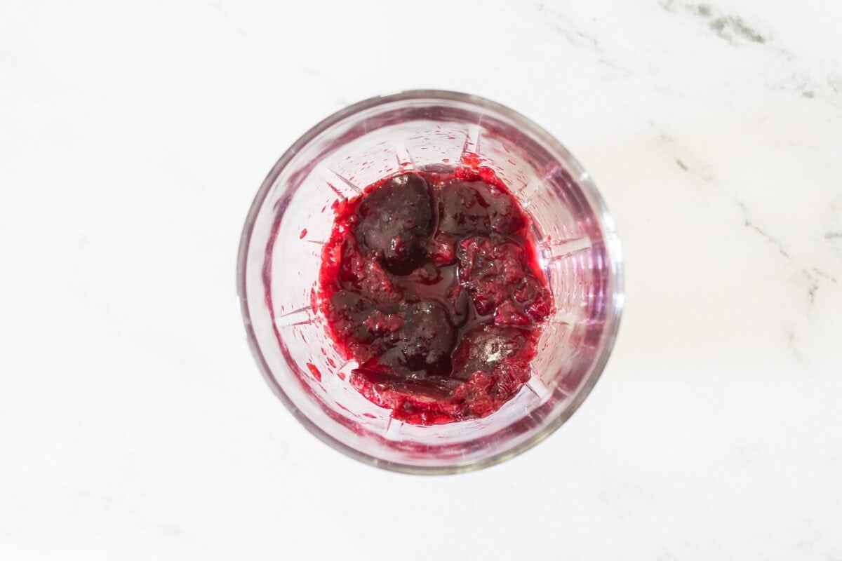 A close-up shot of fresh dark sweet cherries being crushed at the bottom of a lowball glass with a wooden cocktail muddler, releasing rich red juices on a marble countertop.