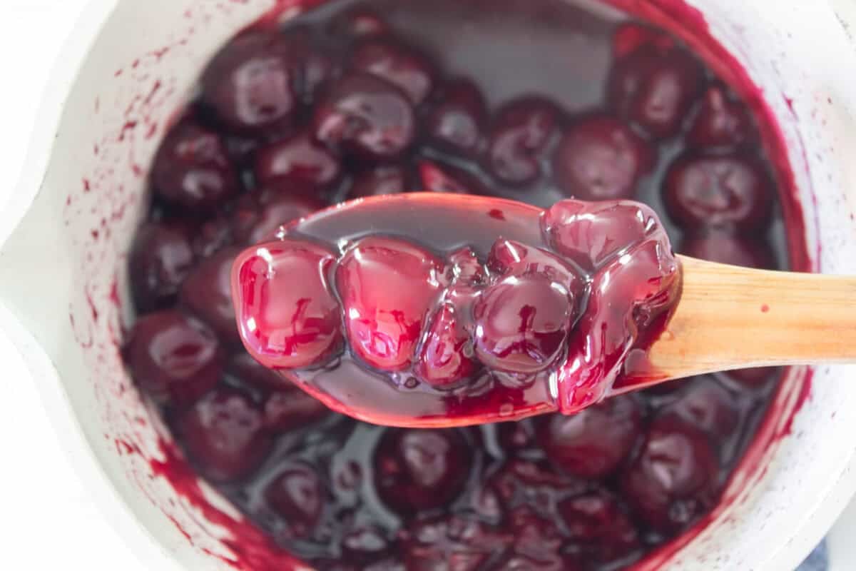 A wooden spoon lifting a scoop of thick, glossy, and dark red cherry turnover filling made from simmered cherries and sweetener out of a white saucepan, showcasing its jammy texture.