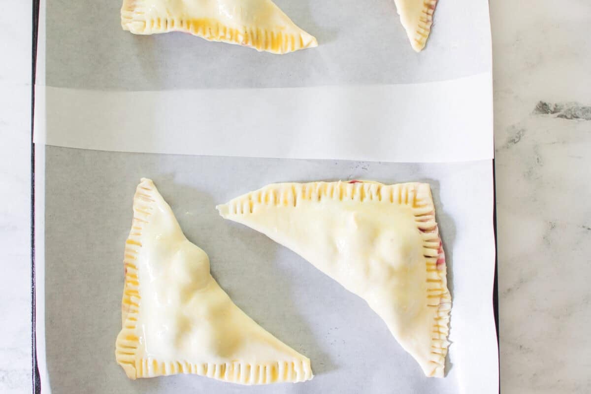 An overhead shot of uncooked, triangular cherry turnovers arranged on a parchment-lined baking sheet, showing the edges neatly crimped with fork tines and the pastry surfaces brushed with a glossy egg wash before baking.