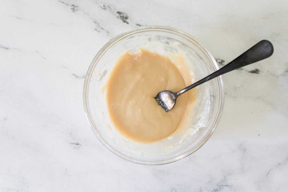 An overhead shot of a clear glass bowl on a marble surface containing a smooth, creamy vanilla glaze being mixed with a metal fork for drizzling over warm cherry turnovers.