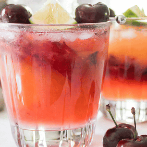 2 glasses of Cherry Mango Margarita on white counter surrounded by a few dark sweet cherries