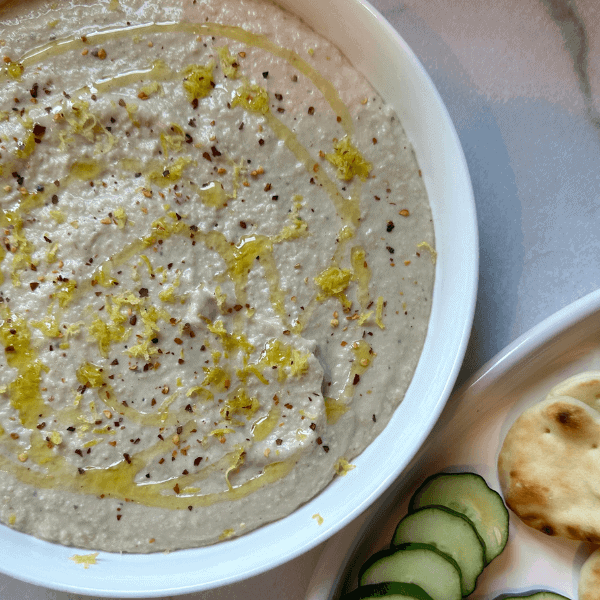 White Bean artichoke dip in a white bowl with a dipper plate of pita bread and cucumbers to the right