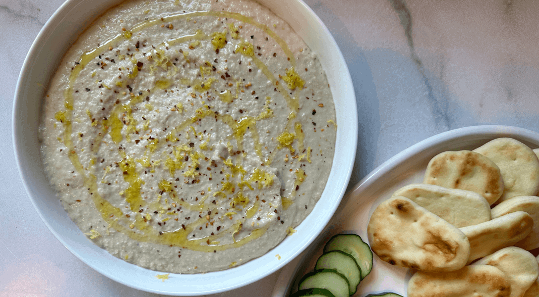 White Bean artichoke dip in a white bowl with a dipper plate of pita bread and cucumbers to the right