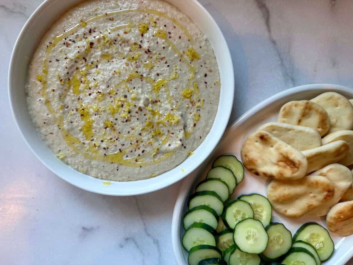 White Bean Artichoke Dip in White bowl with cucumbers and pita bread dippers to the side