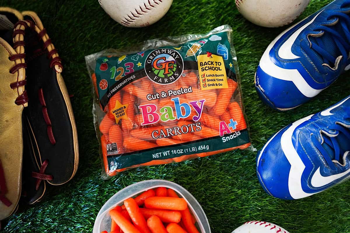 Bag of back to school carrots on grass with soccer cleats, baseballs and a baseball glove