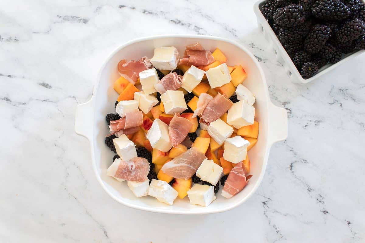 Overhead view of peach and blackberry baked brie with melted brie, prosciutto, fresh peaches, blackberries, and herbs in a white baking dish.