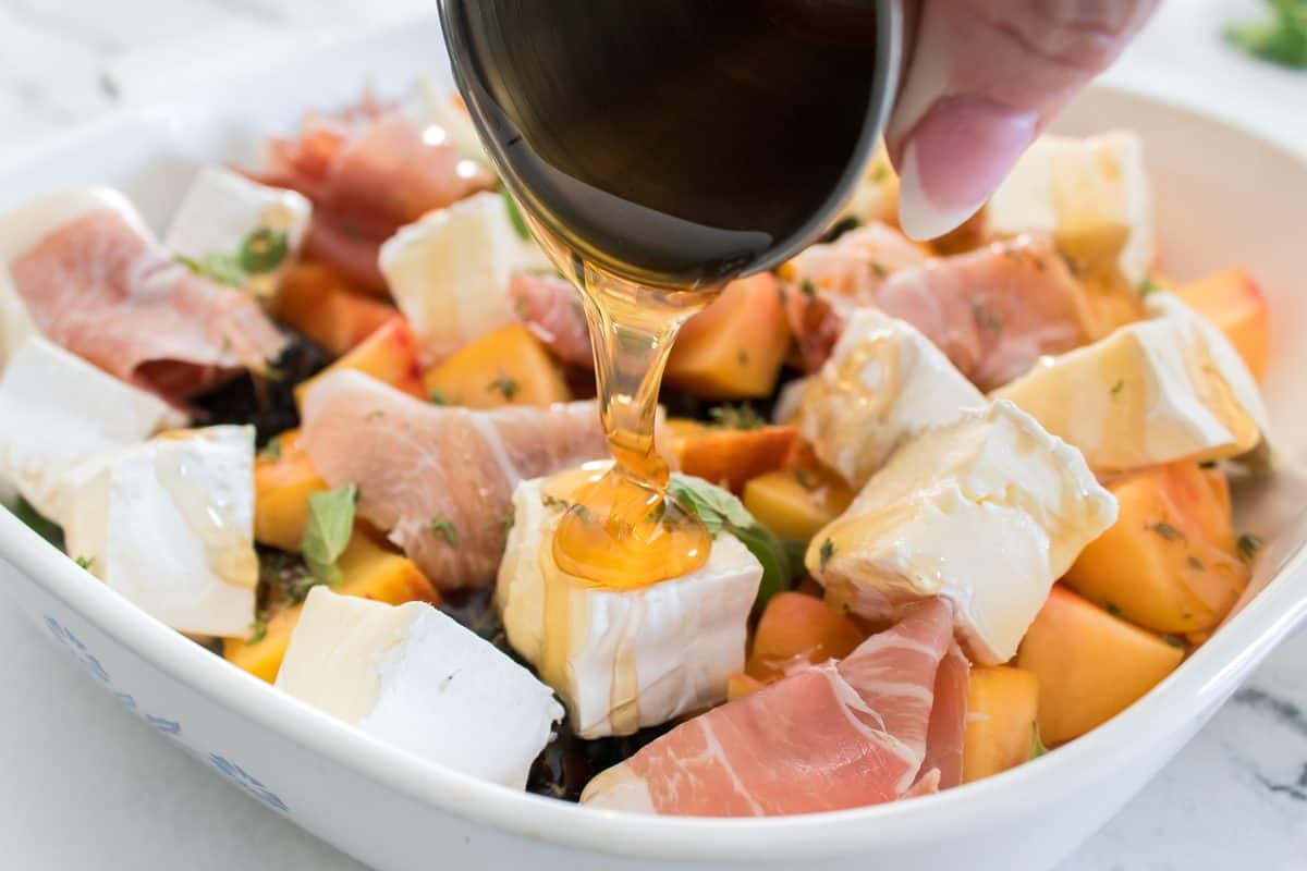 Hot honey being drizzled over peach and blackberry baked brie with prosciutto and fresh herbs in a white baking dish.