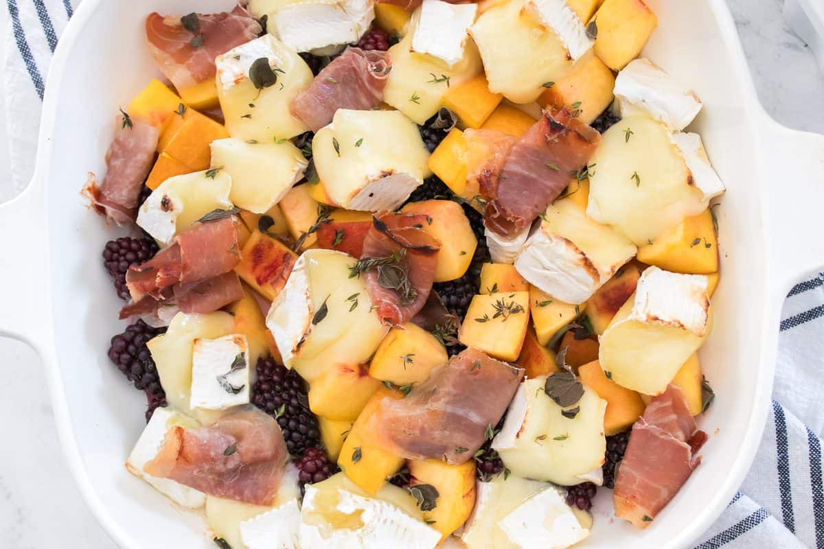 Peach and blackberry baked brie with melted cheese, prosciutto, fresh herbs, and hot honey in a white baking dish.
