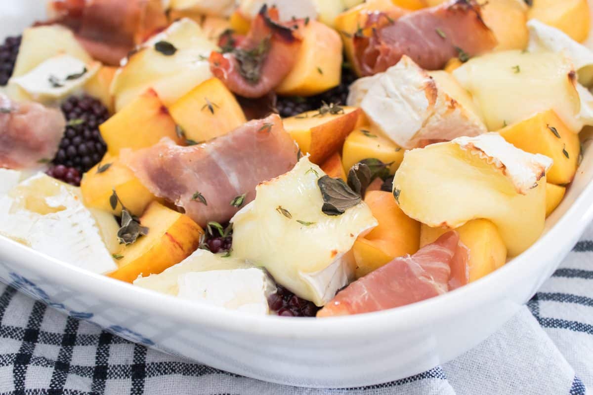 Baked brie with fresh peaches, blackberries, prosciutto, and herbs in a white baking dish, ready to serve as a sweet and savory appetizer.