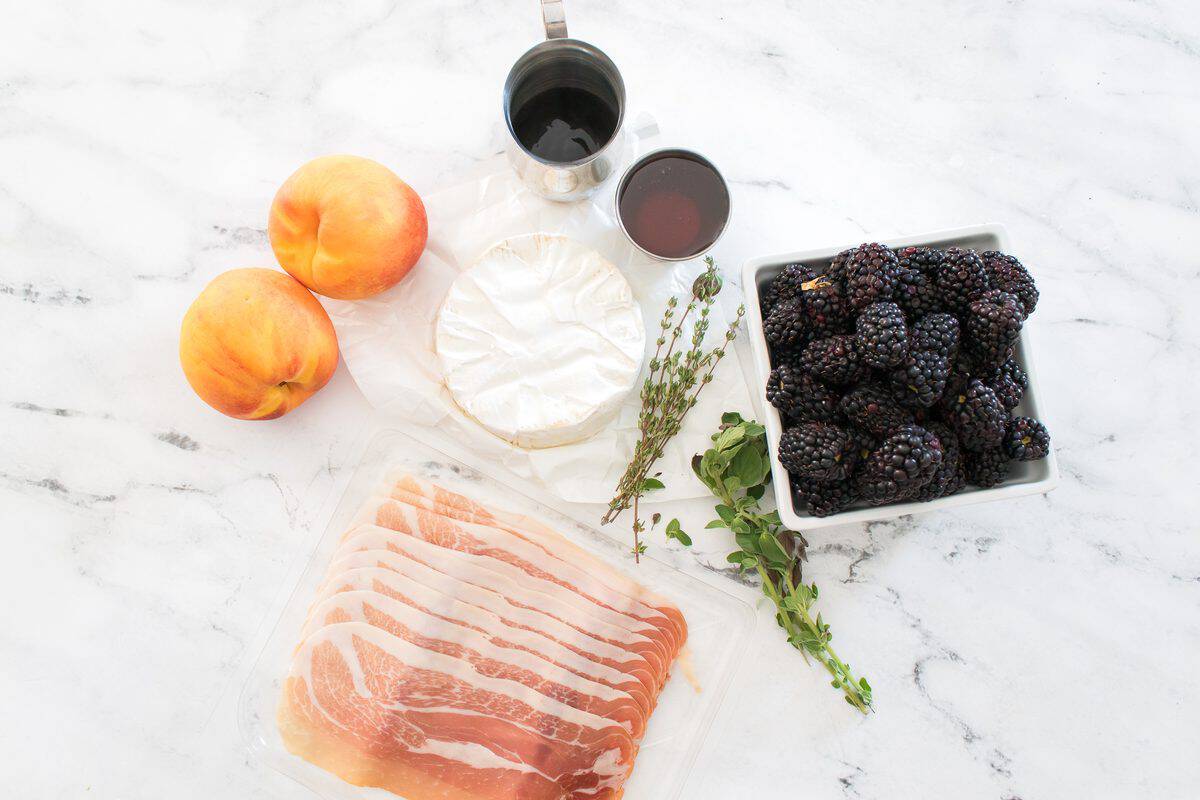 Ingredients for peach and blackberry baked brie arranged on a marble counter, including fresh peaches, blackberries, brie, prosciutto, herbs, honey, and hot honey.