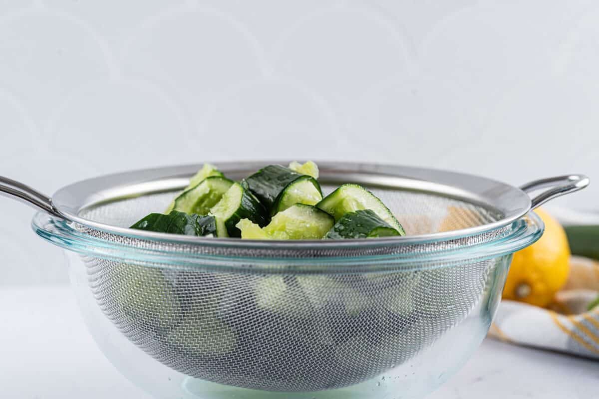 Crushed and sliced cucumber draining in a colander over a bowl.