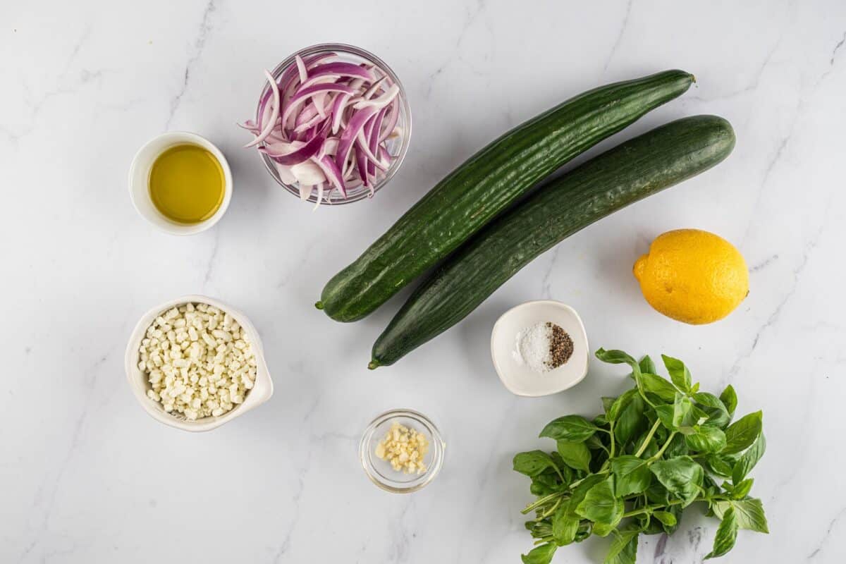 Long English cucumbers, feta, basil, red onion, olive oil, lemon juice, garlic, salt, pepper, and red pepper flakes with a marble bakground.