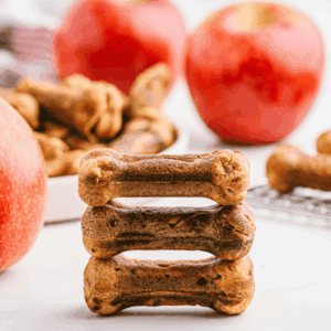 3 apple and bacon dog treats stacked with more treats and apples in background