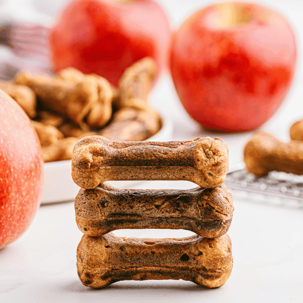 3 apple and bacon dog treats stacked with more treats and apples in background