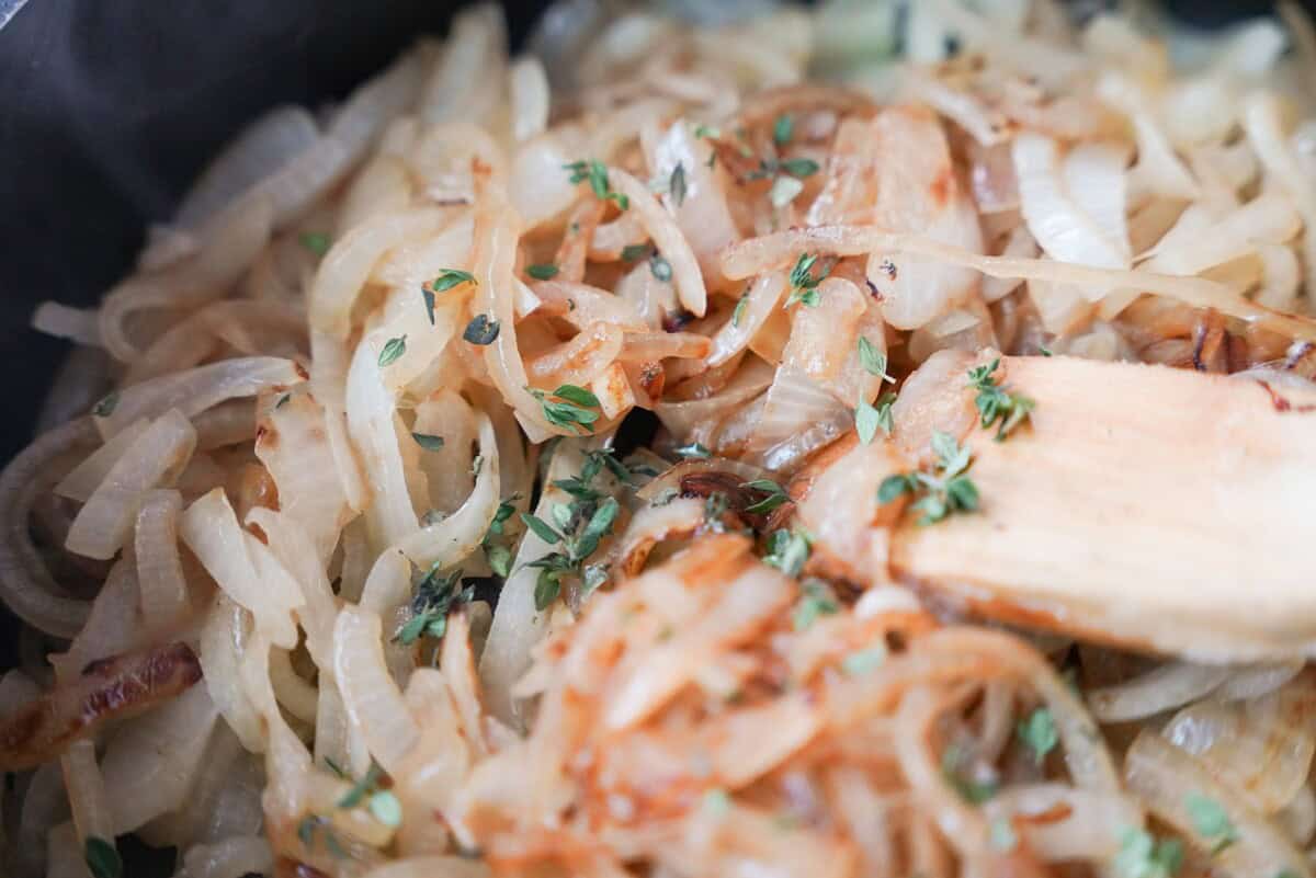 Onion slices cooking in a skillet with fresh thyme, mixing in the beef broth.