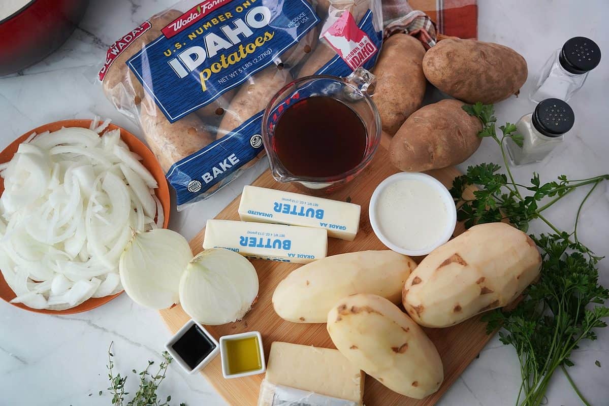 Potatoes, onions, butter, heavy whipping cream, Gruyere cheese, olive oil, Worcestershire sauce, parsley, salt, and pepper on a cutting board