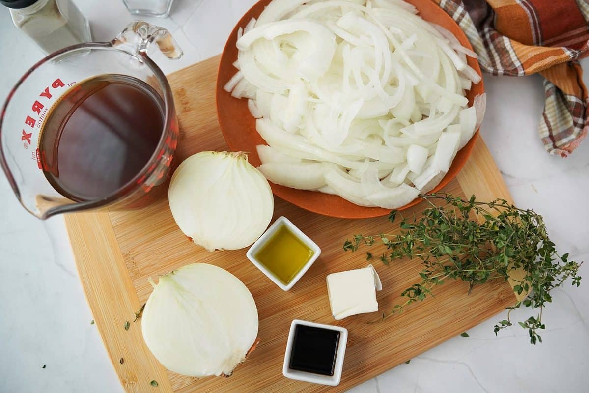 Sliced onions in a bowl on a cutting board with butter, beef broth, thyme,, olive oil, and Worcestershire sauce